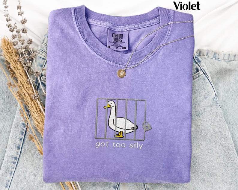 May include: A violet Comfort Colors t-shirt with a graphic of a duck in a cage, a padlock, and the text "got too silly." A silver necklace with a pendant is on the shirt.