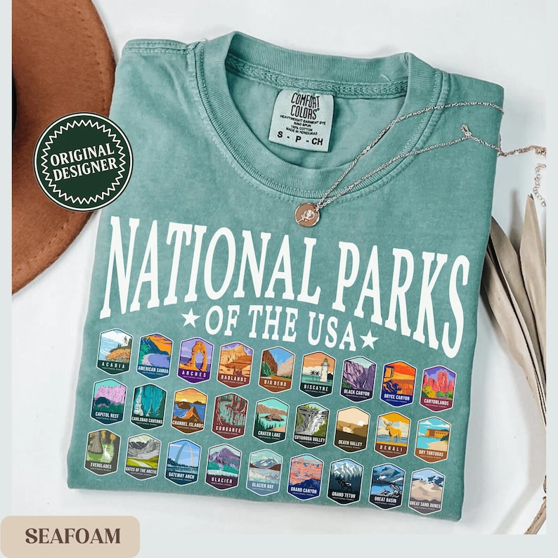 以下が含まれることがあります： シフォングリーンのTシャツに、白い文字で「National Parks of the USA」と書いてあります。Tシャツには、40種類の異なる国立公園のロゴが描かれています。