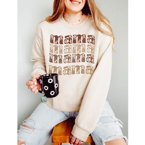 Pode incluir: Uma camisola bege com a palavra "mama" repetida quatro vezes em letras castanhas e bege com detalhes florais. A camisola est&aacute; a ser usada por uma pessoa que segura uma caneca preta com flores brancas.