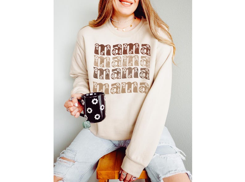 Pode incluir: Uma camisola bege com a palavra "mama" repetida quatro vezes em letras castanhas e bege com detalhes florais. A camisola est&aacute; a ser usada por uma pessoa que segura uma caneca preta com flores brancas.