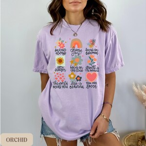 Pu&ograve; includere: Una maglietta color lavanda con un design floreale e il testo "be kind always, choose joy, keep on blooming, you matter, focus on the good, growth is progress, the world needs you, life is beautiful, you are loved".