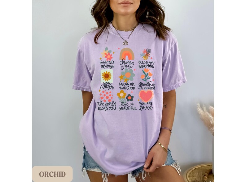 Pu&ograve; includere: Una maglietta color lavanda con un design floreale e il testo "be kind always, choose joy, keep on blooming, you matter, focus on the good, growth is progress, the world needs you, life is beautiful, you are loved".