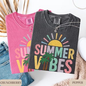 K&ouml;nnte beinhalten: Zwei Comfort Colors T-Shirts, eines in Crunchberry-Pink und eines in Pepper-Grau, mit einem "Summer Vibes"-Grafikdesign. Jedes Shirt hat einen Rundhalsausschnitt und kurze &Auml;rmel. An jedem Shirt befindet sich eine silberne Halskette mit Anh&auml;nger. Die Shirts liegen neben einer Jeans und einem wei&szlig;en Schuh.