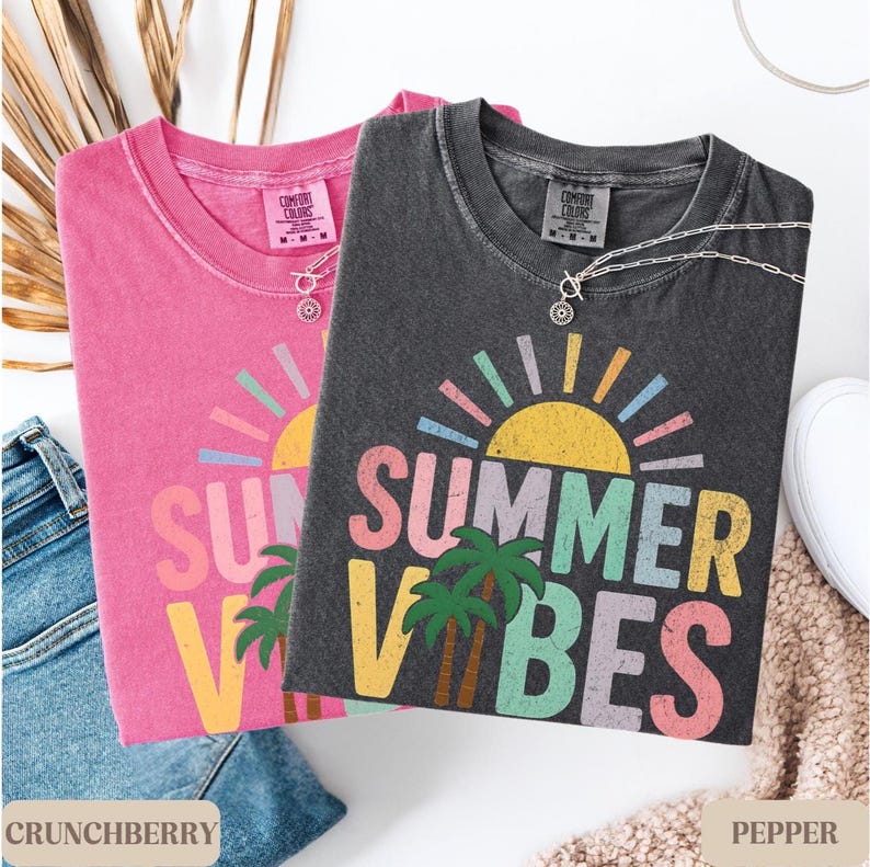 K&ouml;nnte beinhalten: Zwei Comfort Colors T-Shirts, eines in Crunchberry-Pink und eines in Pepper-Grau, mit einem "Summer Vibes"-Grafikdesign. Jedes Shirt hat einen Rundhalsausschnitt und kurze &Auml;rmel. An jedem Shirt befindet sich eine silberne Halskette mit Anh&auml;nger. Die Shirts liegen neben einer Jeans und einem wei&szlig;en Schuh.