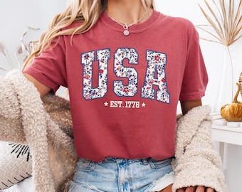May include: A red t-shirt with a white floral print of the word "USA" and the text "EST. 1776" underneath.