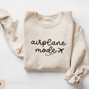 Puede incluir: Una sudadera beige con el texto "airplane mode" y un peque&ntilde;o avi&oacute;n impreso en negro.