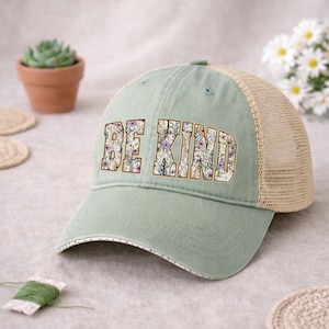 May include: A sage green trucker hat with the words "BE KIND" in a floral design. The hat has a beige mesh back and a curved brim with a dotted trim. A small succulent plant in a terracotta pot is visible in the background.