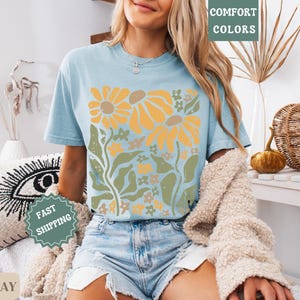 May include: A light blue short-sleeve t-shirt featuring a graphic print of yellow sunflowers with green stems and leaves. The shirt has a crew neckline and is worn with light blue denim shorts and a beige textured cardigan draped over the shoulders.