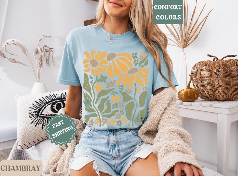 May include: A light blue short-sleeve t-shirt featuring a graphic print of yellow sunflowers with green stems and leaves. The shirt has a crew neckline and is worn with light blue denim shorts and a beige textured cardigan draped over the shoulders.
