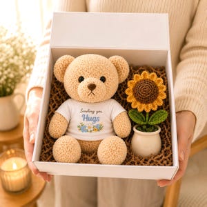 May include: A beige crocheted teddy bear wearing a white t-shirt with the text Sending you Hugs and a floral graphic, placed in a white gift box next to a crocheted sunflower in a small white ceramic pot, surrounded by brown paper filler.