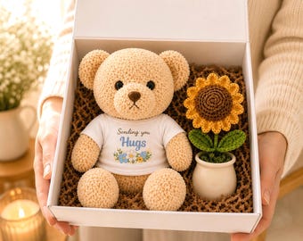 Op de afbeelding: Een beige gehaakte teddybeer in een wit t-shirt met de tekst Sending you Hugs en een bloemenprint, geplaatst in een witte geschenkdoos naast een gehaakte zonnebloem in een kleine witte keramische pot, omringd door bruin opvulmateriaal.