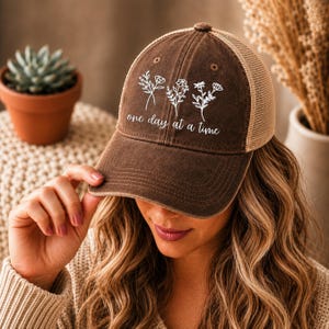 May include: A brown baseball cap with a tan mesh back. The cap features white floral designs and the phrase "one day at a time." The hat is worn by a person.