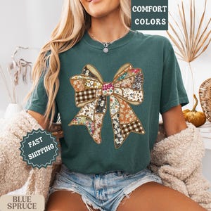 May include: Teal-colored t-shirt featuring a large bow design made of various floral and plaid patterns. The words "COMFORT COLORS" are printed at the top right. The shirt is part of a fall-themed collection.
