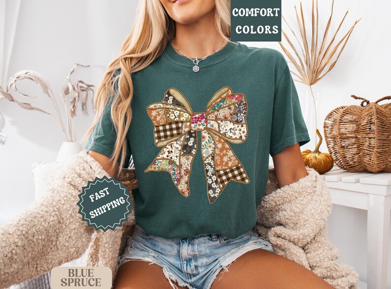 May include: Teal-colored t-shirt featuring a large bow design made of various floral and plaid patterns. The words "COMFORT COLORS" are printed at the top right. The shirt is part of a fall-themed collection.