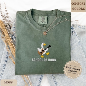 May include: A moss green t-shirt with an embroidered design of a duck playing a guitar. The text "SCHOOL OF HONK" is below the duck. The shirt is a Comfort Colors brand, with an embroidered design.