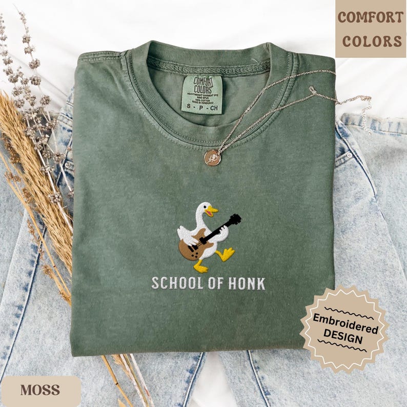 May include: A moss green t-shirt with an embroidered design of a duck playing a guitar. The text "SCHOOL OF HONK" is below the duck. The shirt is a Comfort Colors brand, with an embroidered design.