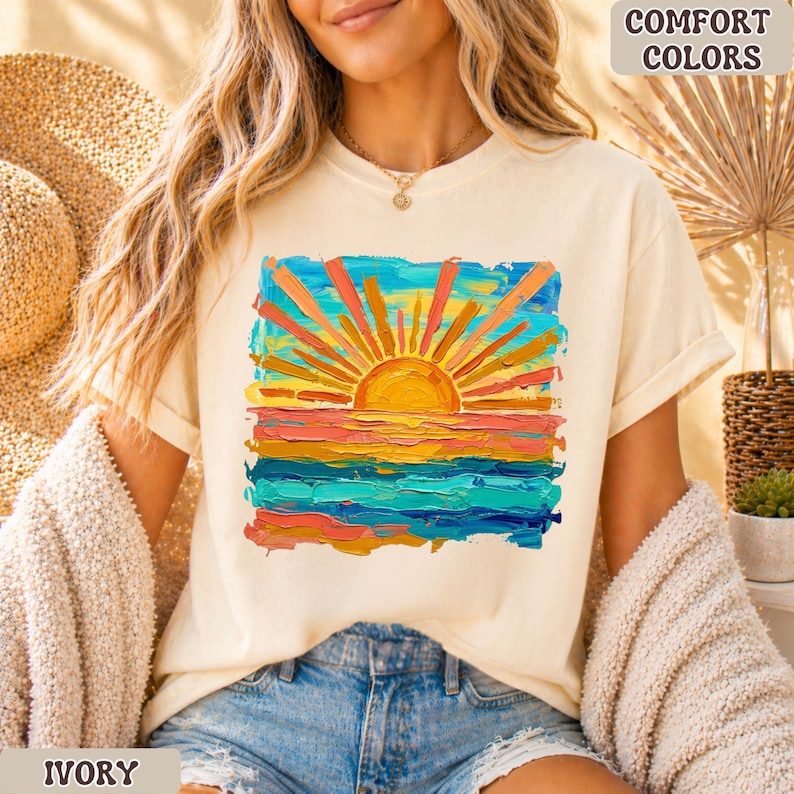 Puede incluir: Camiseta de color marfil con un dise&ntilde;o vibrante de puesta de sol. El gr&aacute;fico representa un sol con rayos naranjas y amarillos sobre un oc&eacute;ano colorido. Las palabras "COMFORT COLORS" y "IVORY" est&aacute;n impresas en la camiseta.