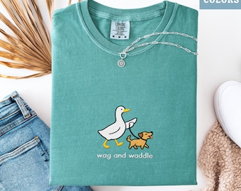 May include: Seafoam green Comfort Colors t-shirt with a graphic of a duck walking a dog, with the text "wag and waddle". A silver necklace is draped on the shirt. The shirt is folded next to a pair of blue jeans, a white shoe, and a beige blanket.