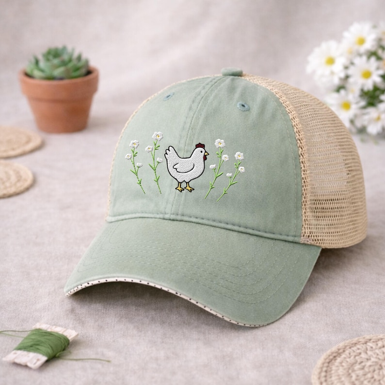 May include: A sage green trucker hat with a white embroidered chicken and floral design. The hat features a mesh back, a curved brim, and a button on top. The brim is trimmed with small white dots.