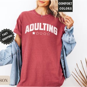 May include: Crimson-colored t-shirt with the word "ADULTING" in white block letters, above four white stars. The shirt is worn with a denim jacket. The words "COMFORT COLORS" and "FAST SHIPPING" are also visible.