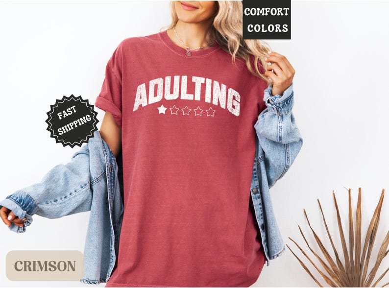 May include: Crimson-colored t-shirt with the word "ADULTING" in white block letters, above four white stars. The shirt is worn with a denim jacket. The words "COMFORT COLORS" and "FAST SHIPPING" are also visible.