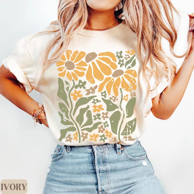 May include: An ivory short-sleeve t-shirt featuring a large central graphic print of stylized flowers in shades of mustard yellow, sage green, and muted brown. The floral design has a distressed, textured appearance with various leaf and petal shapes.