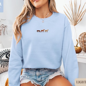 K&ouml;nnte beinhalten: Hellblaues Sweatshirt mit Pferde-Stickerei. Die Worte "SUPER SOFT" befinden sich in einer hellblauen Box oben links. Die Worte "LIGHT BLUE" befinden sich in einer hellbeigen Box unten rechts.