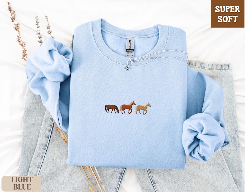 K&ouml;nnte beinhalten: Hellblaues Sweatshirt mit Pferde-Stickerei. Das Sweatshirt hat einen Rundhalsausschnitt und lange &Auml;rmel. Der Text "SUPER SOFT" befindet sich oben rechts. Der Text "LIGHT BLUE" befindet sich unten links.