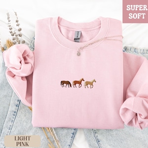 K&ouml;nnte beinhalten: Hellrosa Sweatshirt mit gestickten Pferden. Das Rundhals-Sweatshirt hat die Worte "SUPER SOFT" in einem rosa Feld oben rechts und "LIGHT PINK" in einem beigen Feld unten links. Das Sweatshirt liegt gefaltet auf einer Jeans.