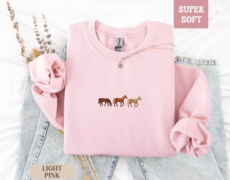 K&ouml;nnte beinhalten: Hellrosa Sweatshirt mit gestickten Pferden. Das Rundhals-Sweatshirt hat die Worte "SUPER SOFT" in einem rosa Feld oben rechts und "LIGHT PINK" in einem beigen Feld unten links. Das Sweatshirt liegt gefaltet auf einer Jeans.