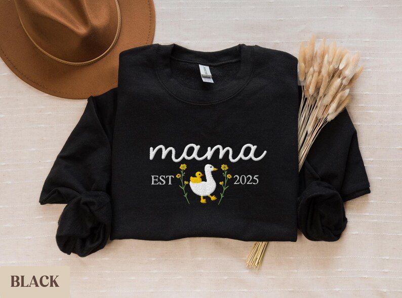 May include: Black sweatshirt with the text "mama est 2025" and an embroidered image of a white duck with a yellow duckling on its back.