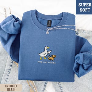 May include: Indigo blue sweatshirt with a crew neck and long sleeves. The front features an embroidered design of a white duck walking a brown dog, with the words "wag and waddle" below. The upper right corner has a "SUPER SOFT" label.