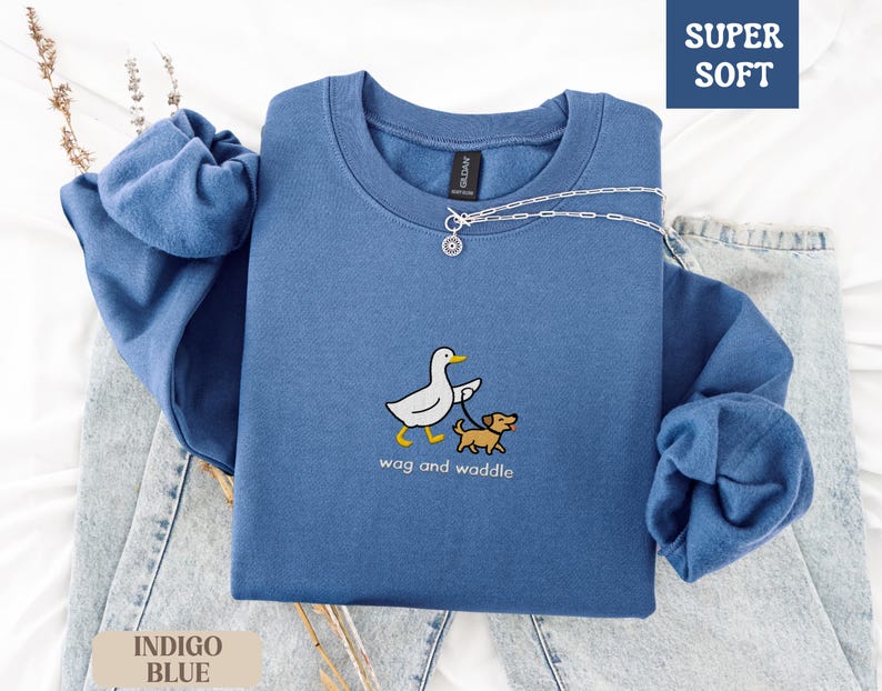 May include: Indigo blue sweatshirt with a crew neck and long sleeves. The front features an embroidered design of a white duck walking a brown dog, with the words "wag and waddle" below. The upper right corner has a "SUPER SOFT" label.