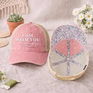 May include: Pink trucker hat with the text "I AM WITH YOU always" in white script. The hat has a mesh back and a floral patterned interior. The hat is displayed next to another hat with the interior visible.