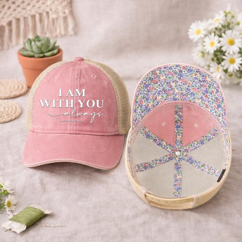 May include: Pink trucker hat with the text "I AM WITH YOU always" in white script. The hat has a mesh back and a floral patterned interior. The hat is displayed next to another hat with the interior visible.