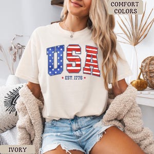 May include: Ivory-colored t-shirt with a patriotic "USA" design. The letters are red and blue, with a star-spangled blue "U" and red and white stripes in the "S" and "A". The text "EST. 1776" is below the letters. The shirt is part of the "Comfort Colors" collection.