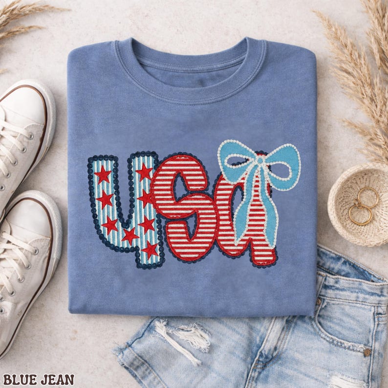 Op de afbeelding: Een lichtblauw t-shirt met het woord "USA" geborduurd in rood, wit en blauw. De "U" heeft rode sterren op een blauwe achtergrond, terwijl de "S" en "A" rood-wit gestreept zijn. Aan de rechterkant zit een lichtblauwe strik.