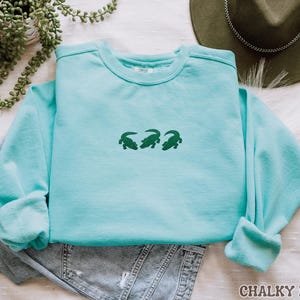 May include: A light blue sweatshirt with three green alligator designs embroidered on the front. The sweatshirt is folded over a pair of denim shorts. The text "CHALKY MINT" is at the bottom right corner of the image.