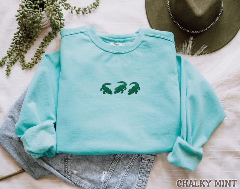 May include: A light blue sweatshirt with three green alligator designs embroidered on the front. The sweatshirt is folded over a pair of denim shorts. The text "CHALKY MINT" is at the bottom right corner of the image.