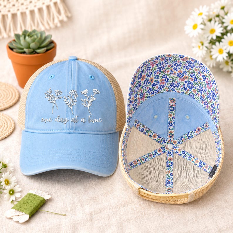 May include: A light blue trucker hat with a floral design and the phrase "one day at a time." The hat has a beige mesh back and a floral patterned interior. The hat is displayed next to a second hat with the interior visible.