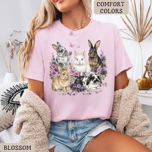Puede incluir: Camiseta rosa claro con un dise&ntilde;o de cinco conejos rodeados de flores moradas y una mariposa. El texto "COMFORT COLORS" est&aacute; en la esquina superior derecha y "BLOSSOM" en la inferior izquierda.
