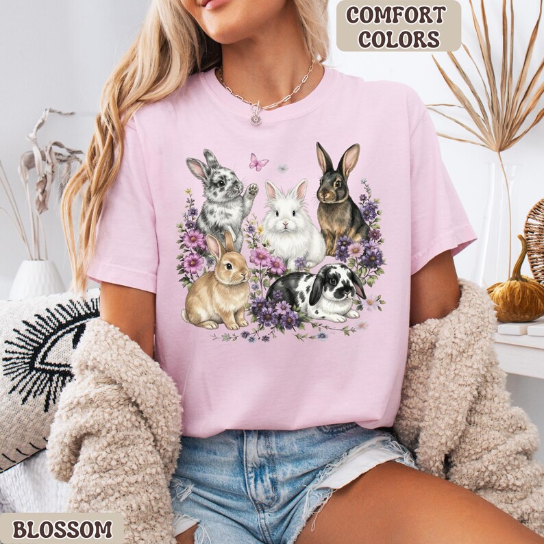 Puede incluir: Camiseta rosa claro con un dise&ntilde;o de cinco conejos rodeados de flores moradas y una mariposa. El texto "COMFORT COLORS" est&aacute; en la esquina superior derecha y "BLOSSOM" en la inferior izquierda.