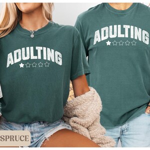 May include: Two people wearing matching blue spruce-colored t-shirts with the word "ADULTING" in white block letters. The word is arched above five white stars. The shirts have short sleeves and a relaxed fit.