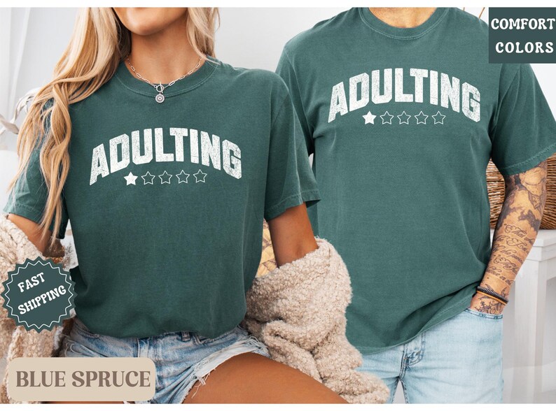May include: Two people wearing matching blue spruce-colored t-shirts with the word "ADULTING" in white block letters. The word is arched above five white stars. The shirts have short sleeves and a relaxed fit.
