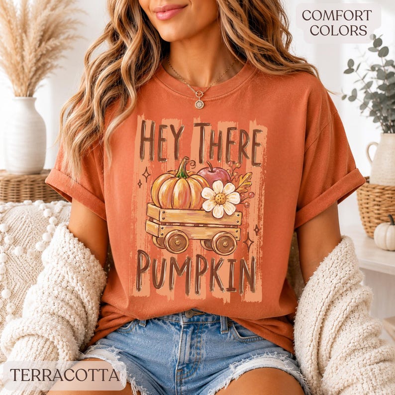 Op de afbeelding: Een terracotta-kleurig t-shirt met korte mouwen en een afbeelding van een houten wagen met een pompoen, een appel en een witte bloem. De tekst HEY THERE PUMPKIN staat in donkerbruine letters boven en onder de illustratie van de wagen.