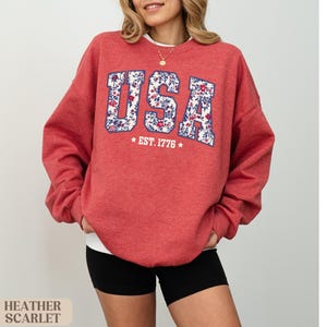 Peut inclure: Sweat-shirt rouge &eacute;carlate avec "USA" en lettres &agrave; imprim&eacute; floral et "EST. 1776" en dessous. Le sweat-shirt &agrave; col rond est associ&eacute; &agrave; un short noir. L'image comprend &eacute;galement le texte "HEATHER SCARLET".