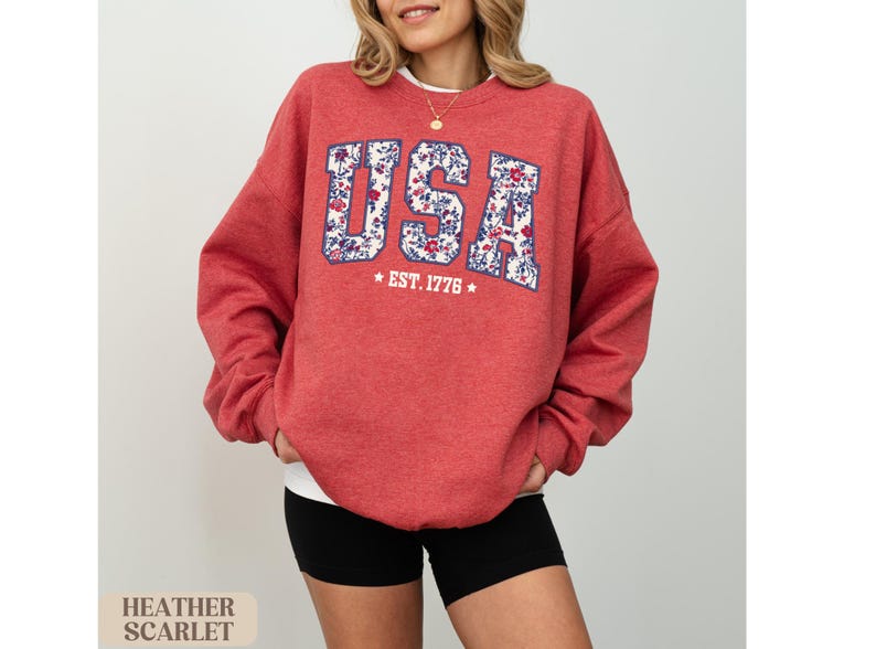 Peut inclure: Sweat-shirt rouge &eacute;carlate avec "USA" en lettres &agrave; imprim&eacute; floral et "EST. 1776" en dessous. Le sweat-shirt &agrave; col rond est associ&eacute; &agrave; un short noir. L'image comprend &eacute;galement le texte "HEATHER SCARLET".