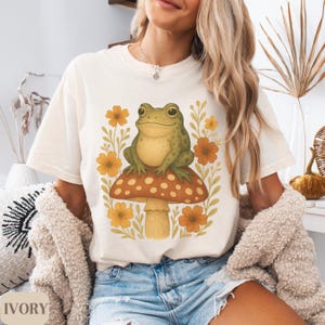 May include: A white t-shirt featuring a green frog perched on a mushroom with yellow and orange flowers. The text "IVORY" is in the bottom left corner of the image.
