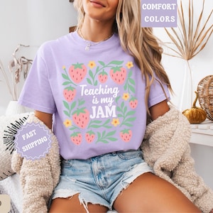 May include: A light purple t-shirt with the text "Teaching is my JAM" surrounded by strawberry and leaf graphics. The shirt is part of the "Comfort Colors" collection and is a casual wear item.