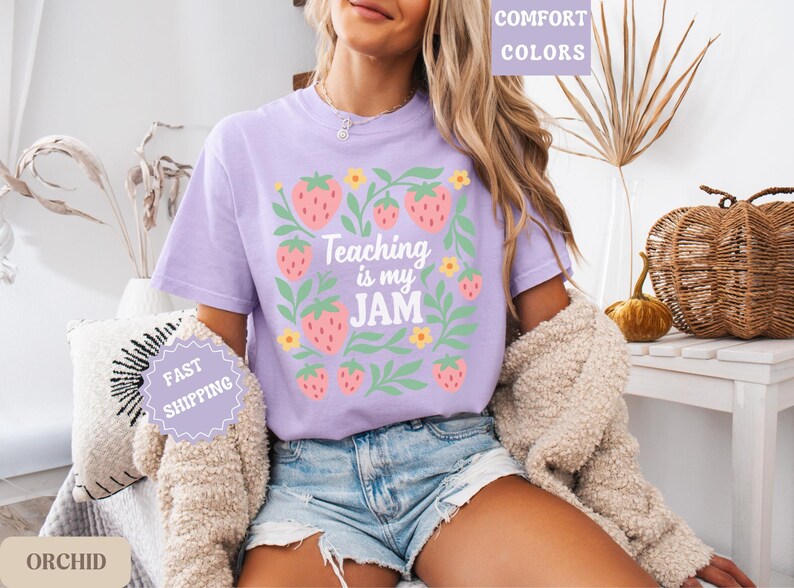 May include: A light purple t-shirt with the text "Teaching is my JAM" surrounded by strawberry and leaf graphics. The shirt is part of the "Comfort Colors" collection and is a casual wear item.